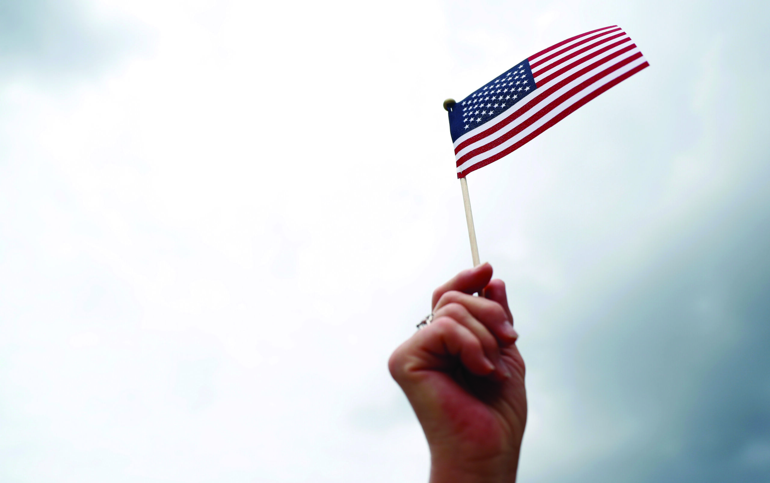 A hand holds a small American flag against the backdrop of a cloudy sky.