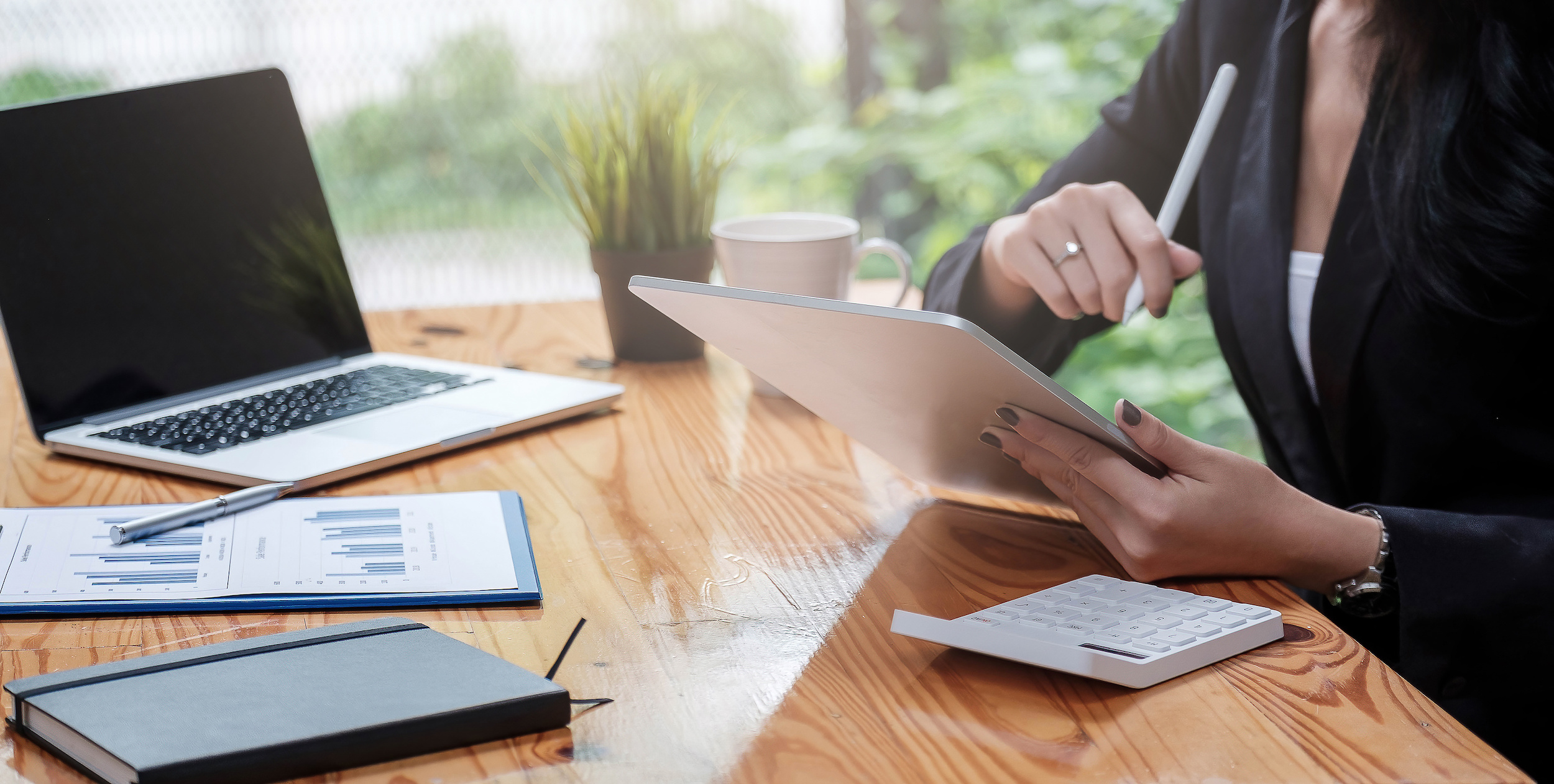 View of a female accountant using a checklist while a chart, laptop, notebook, and calculator lay on the wooden table around her.