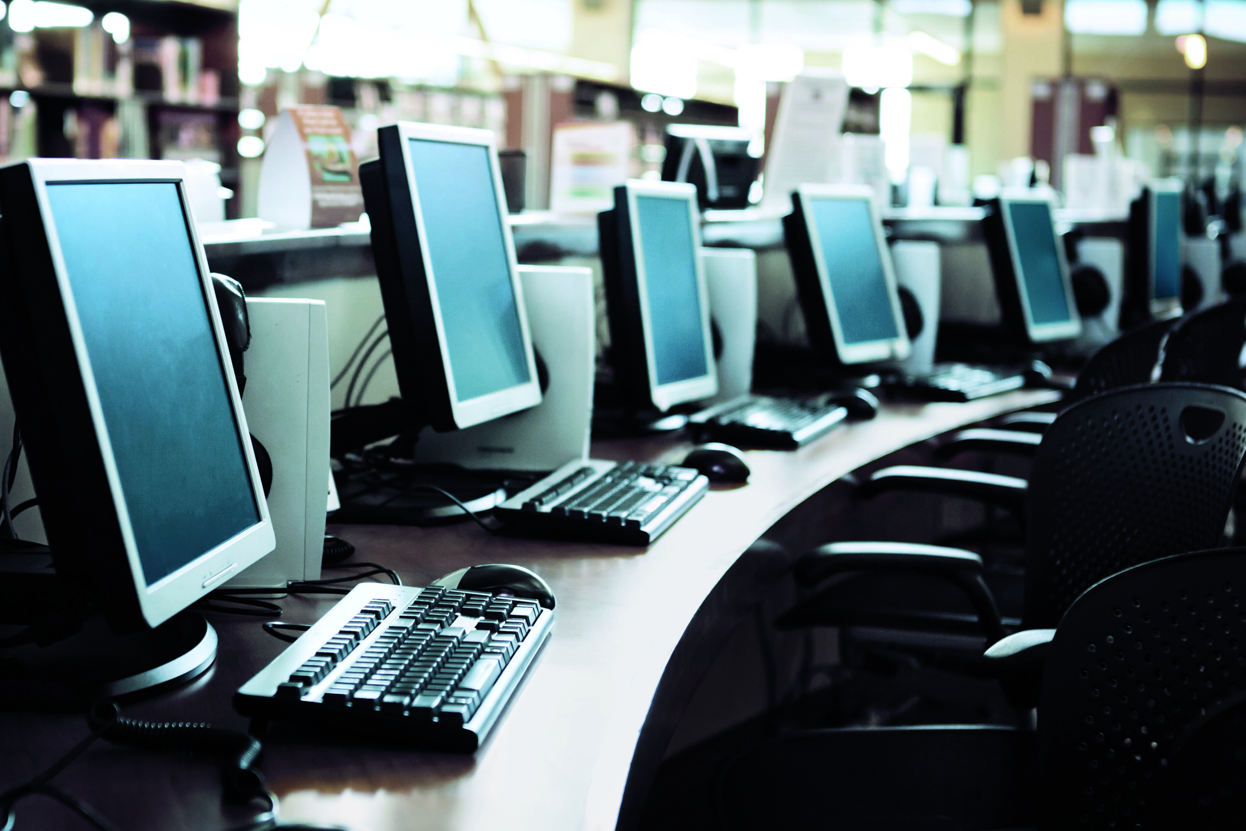 Closeup of a row of desktop computers with keyboards along a continuous desk in a library.