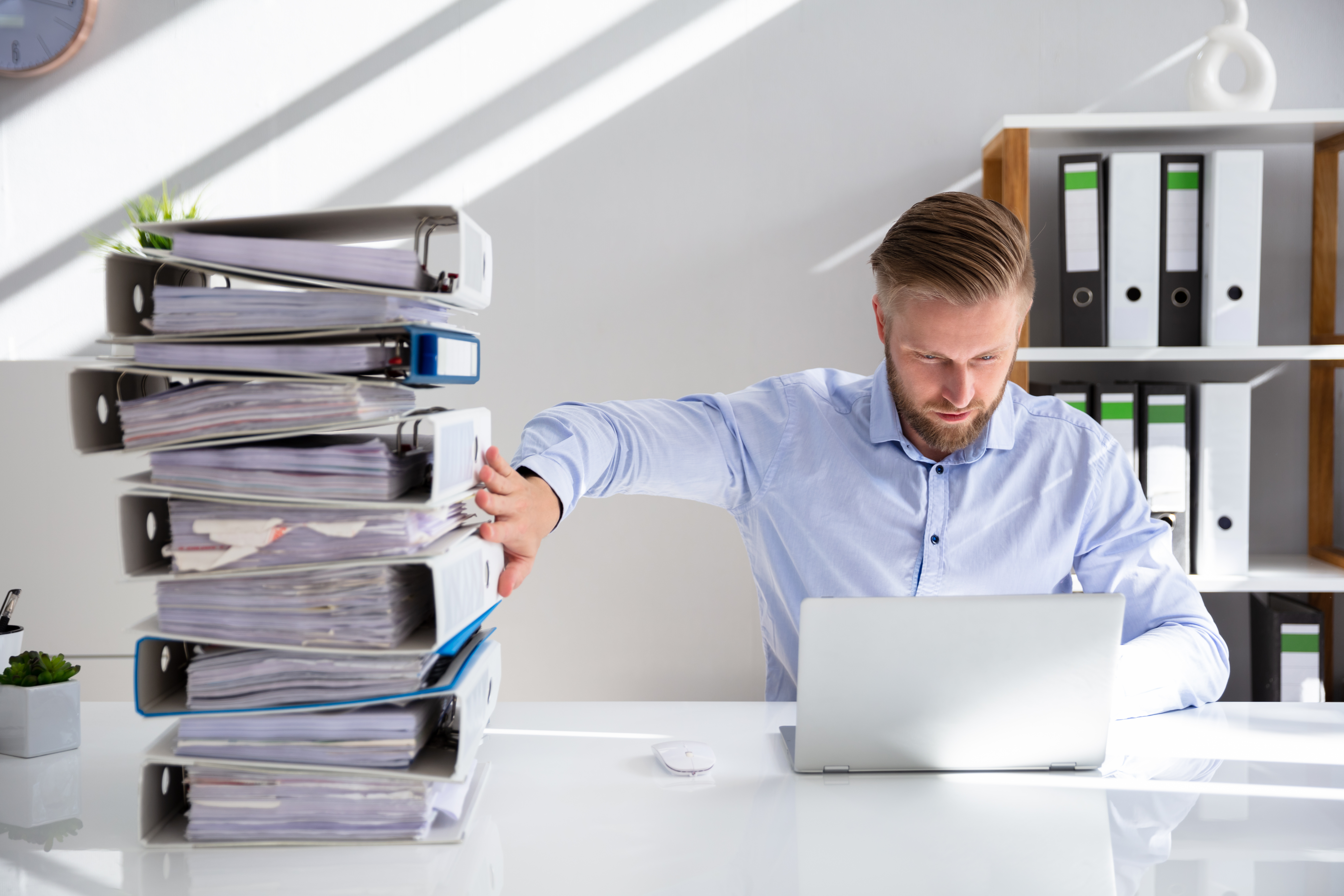 A male accountant working on a laptop pushes a stack of binders full of papers to the side.