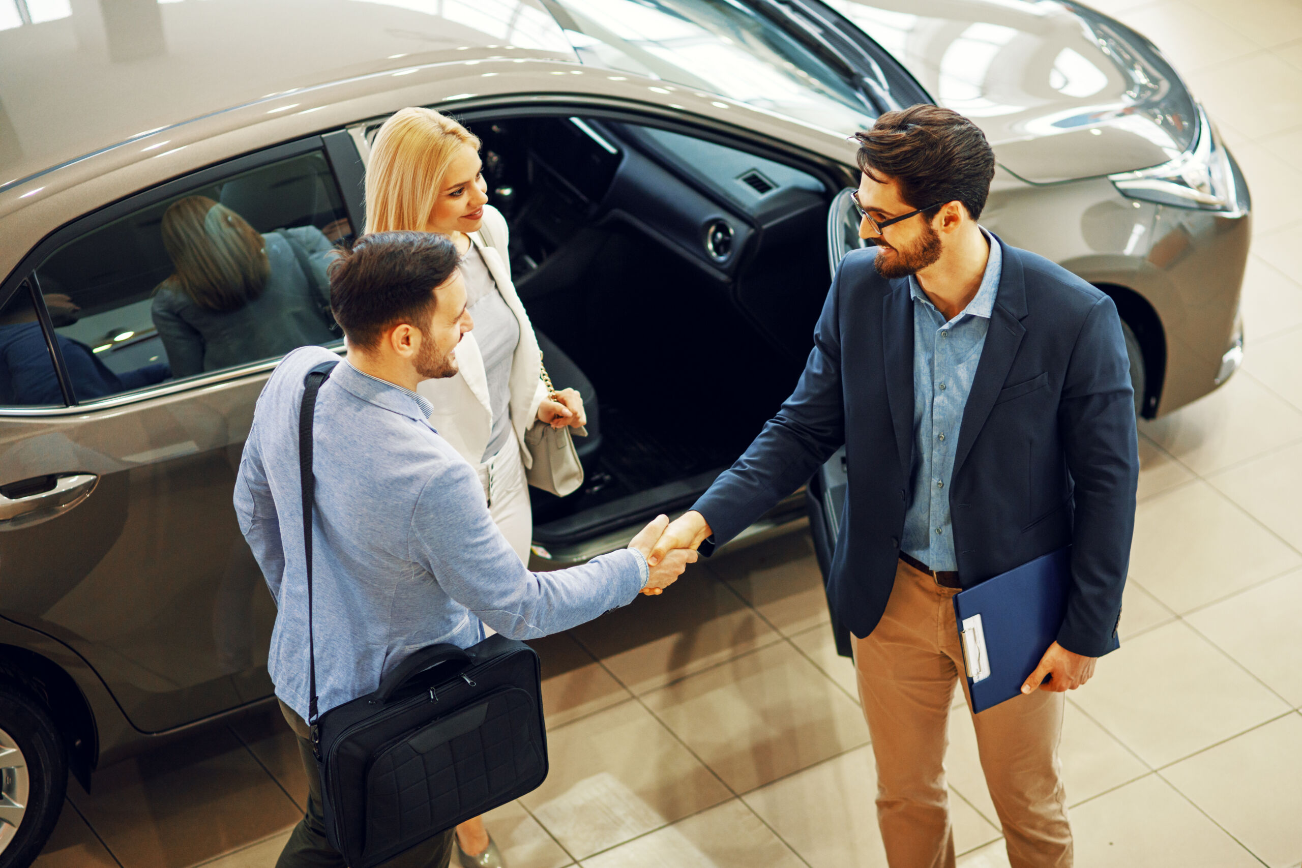 A young couple shakes hands with a car salesman in front a brown sedan.