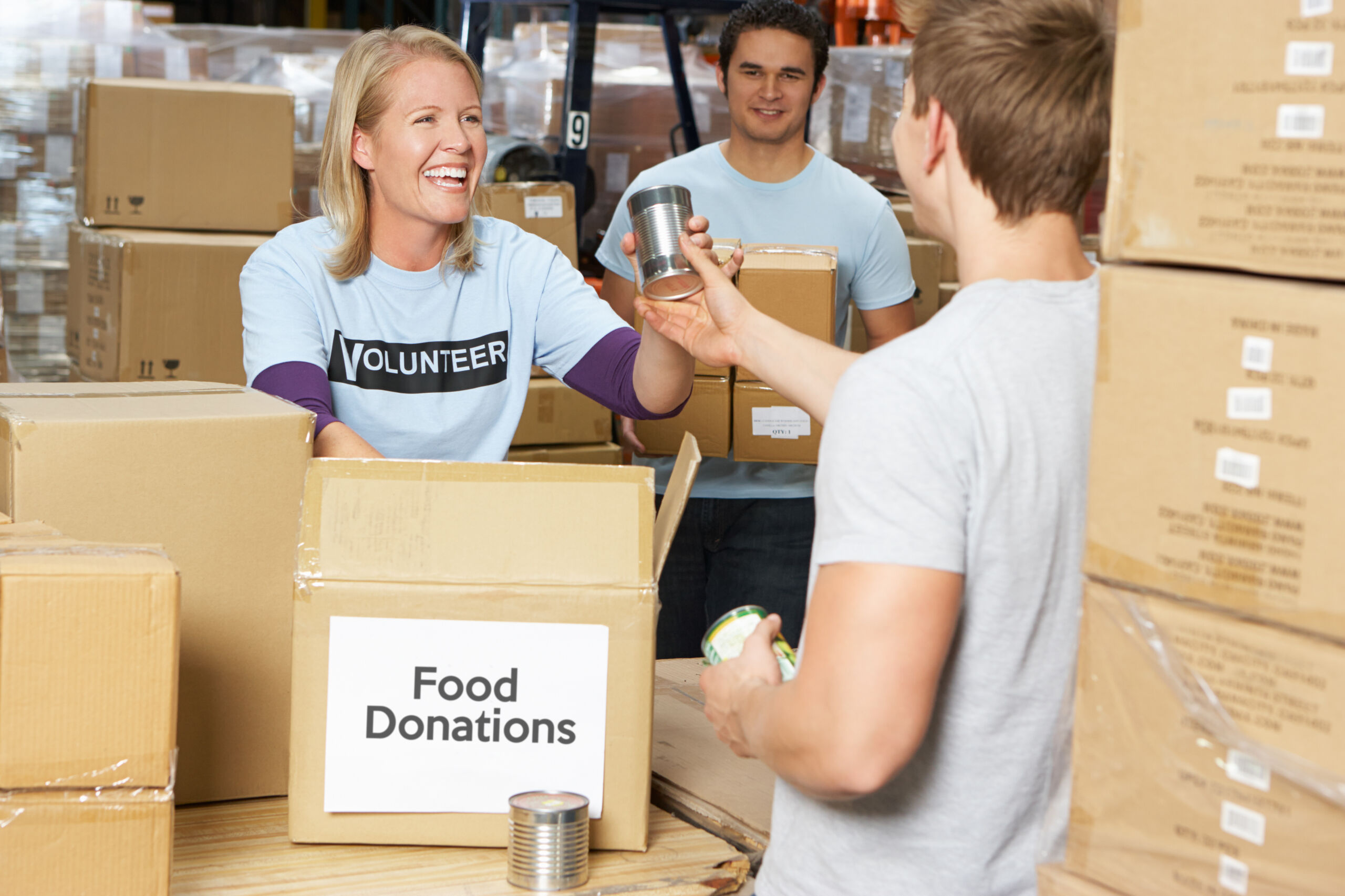 A smiling volunteer standing near piles of boxes receives a can of food to put into a box labeled 'food donations.'