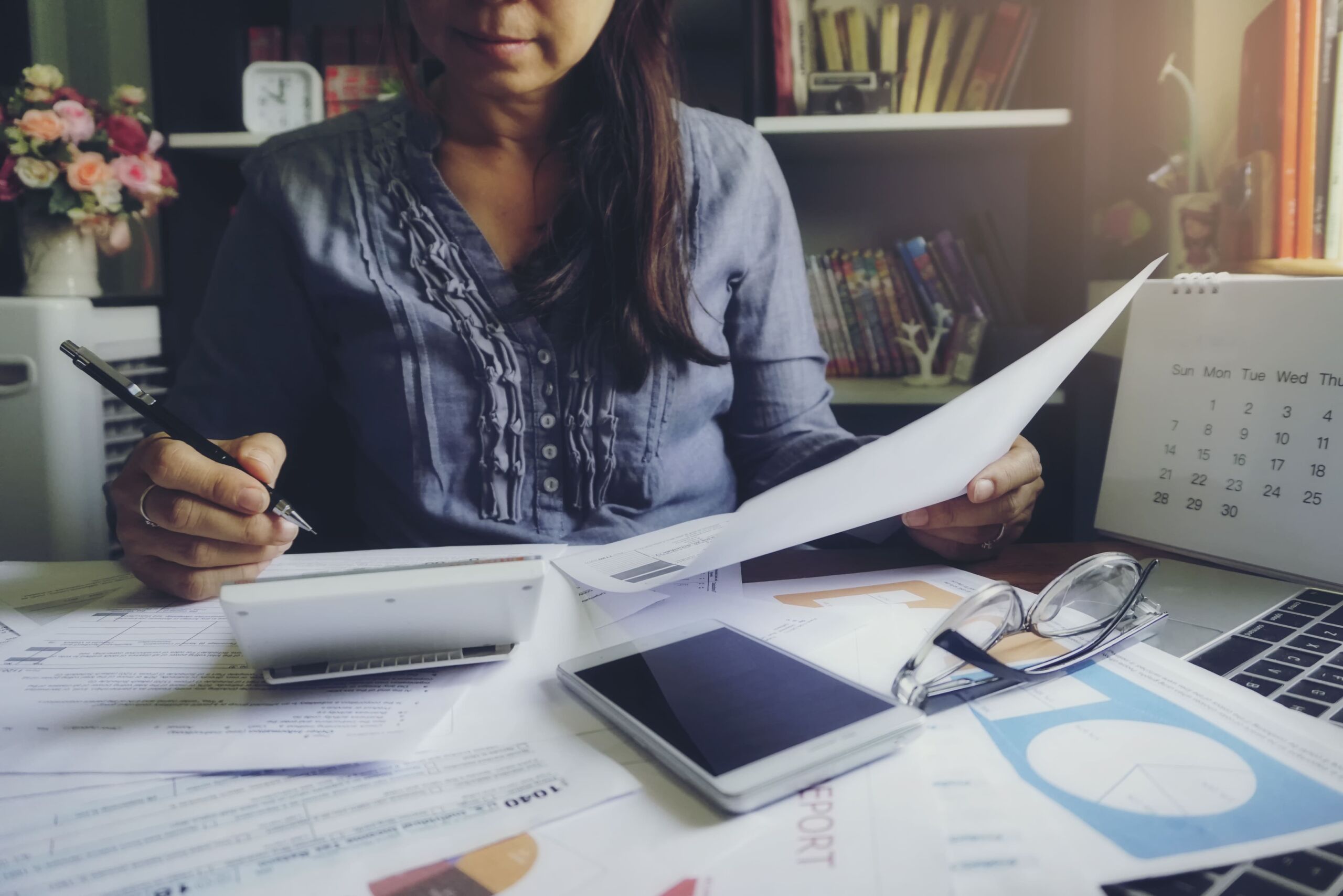 A Black female accountant working at a desk spread out with papers, a laptop, a calendar, a calculator, and a phone.