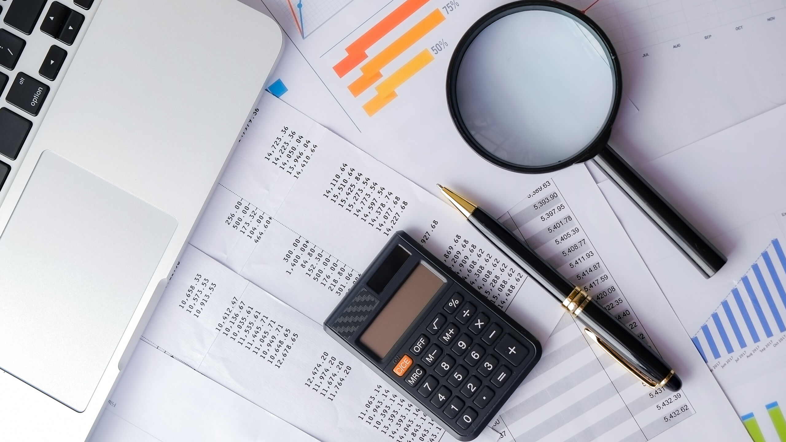 A magnifying glass, pen, calculator, and laptop lay on top of paper financial statements and bar graphs.