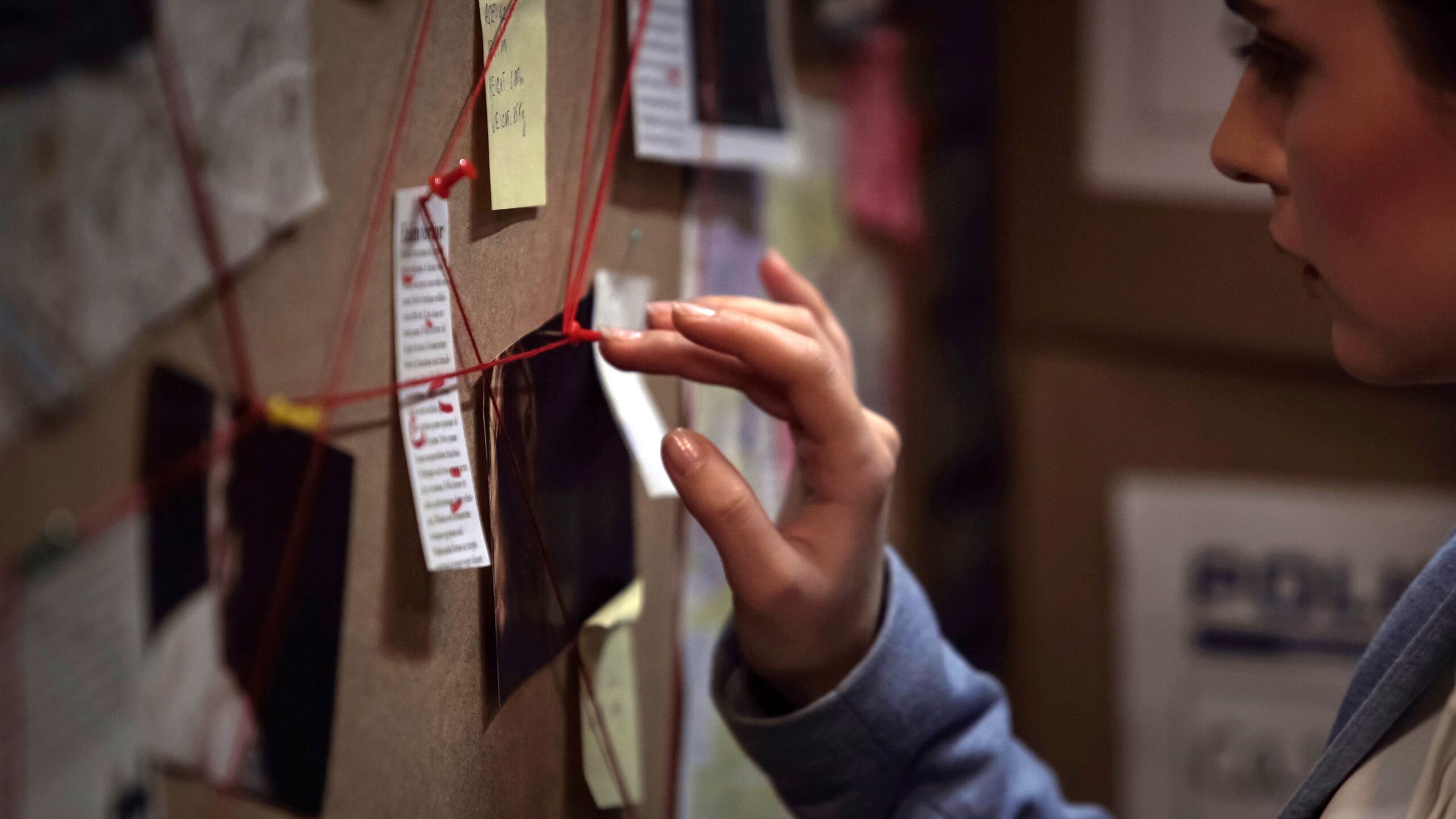 A female investigator looks at a corkboard with red string tying together various pinned up documents.
