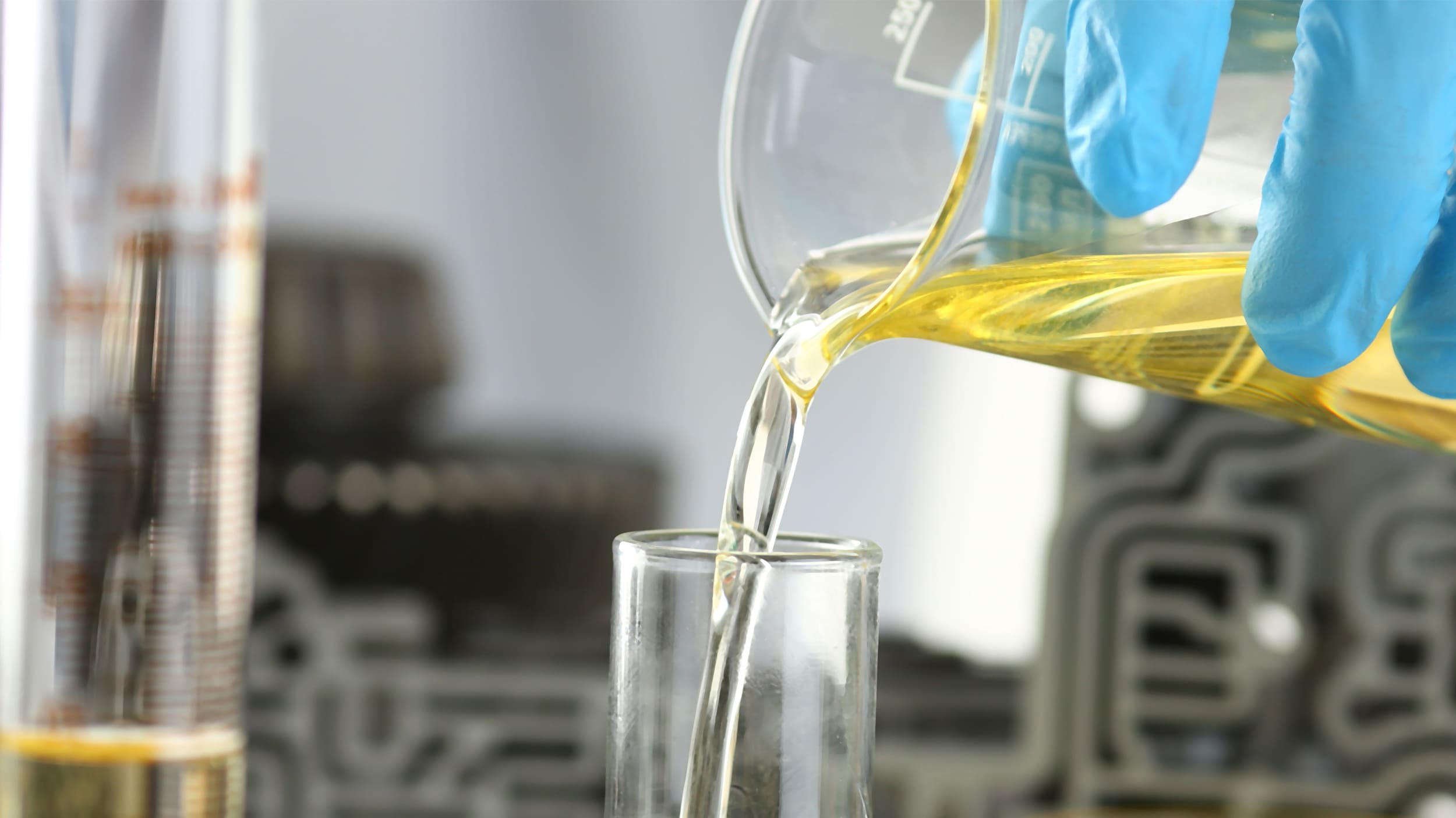 A blue gloved hand pours a yellow chemical into a glass from a beaker.